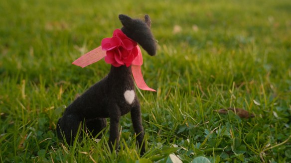 A felted symbol of Lucy, wearing a bright pink fabric flower collar, sitting in the grass at the Schloss Leopoldskron.