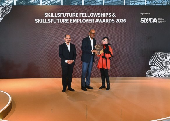Dawn-joy, wearing a bright orange top, black jeans and tall boots, holding a large Lucy plushie, receives her award plaque from President Tharman. On the other side of the President, stands Senior Parliamentary Secretary, Dr Syed Harun Alhabsyi.